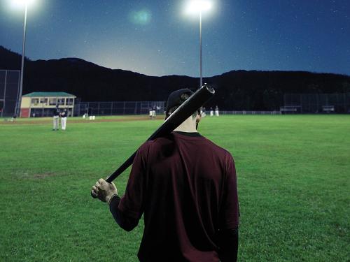 A man on an empty baseball field at night with lights on holds baseball bat over his shoulder.