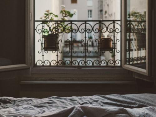 Bed with crumpled sheets in foreground, windows are open with view to outside.