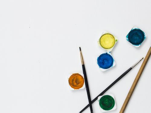 Horizontal photo of paint brushes and yellow, orange, green, and blue water color paints laying on a piece of paper.