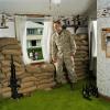 Jeff Gramlich in green carpeted bedroom with machine gun, sandbags, empty beer bottles, looking out at lawn.